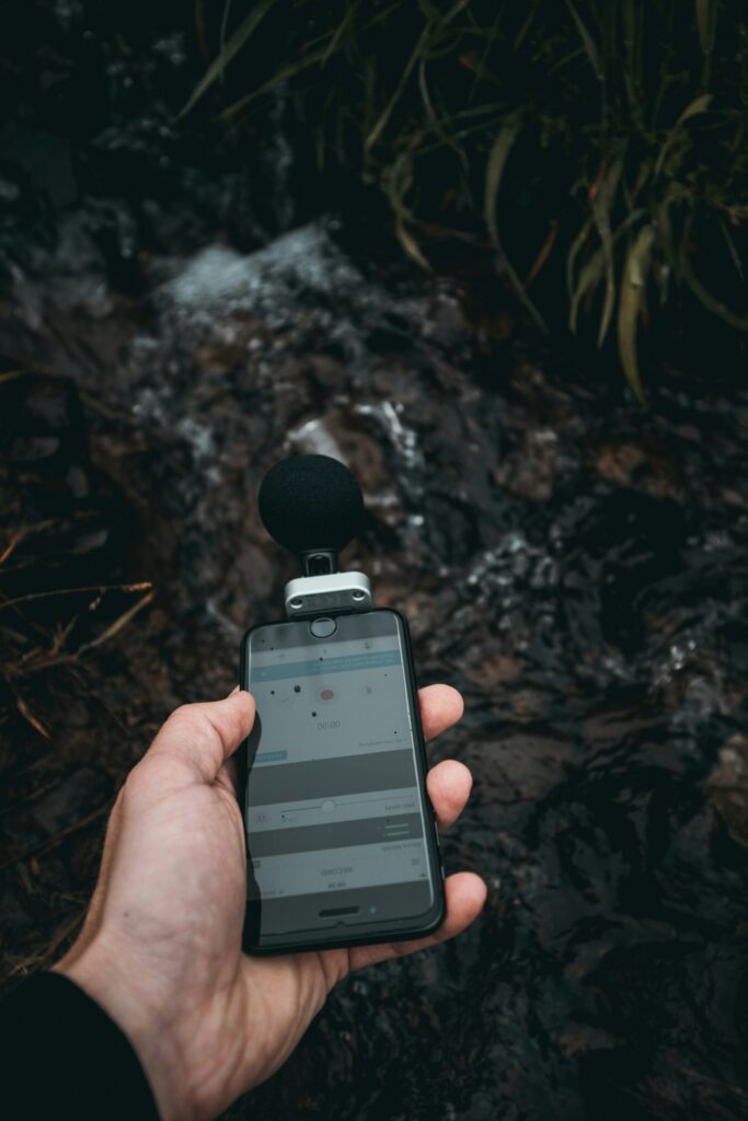 A person holds a phone with a microphone attached to record the sounds of a stream in the forest.